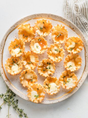 Gold-rimmed speckled plate holding apricot brie phyllo cups garnished with fresh thyme leaves.