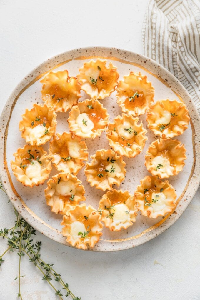 Gold-rimmed speckled plate holding apricot brie phyllo cups garnished with fresh thyme leaves.