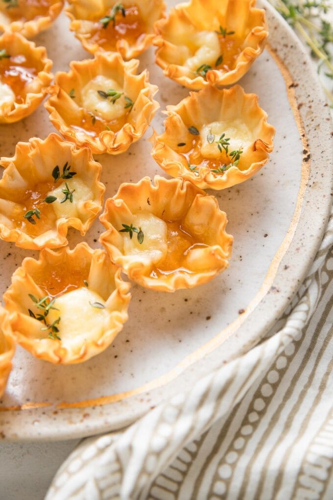 Close up angled view of a miniature phyllo shell filled with melted brie, apricot jam, and a garnish of fresh thyme leaves.