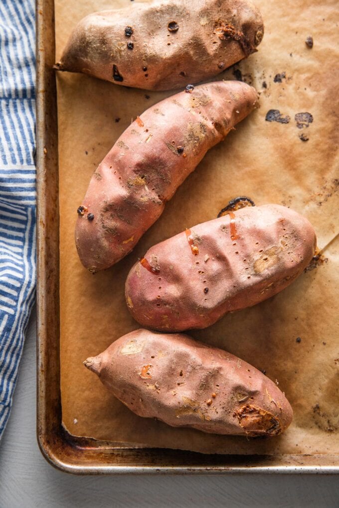 Four tender baked sweet potatoes lying on a sheet pan.