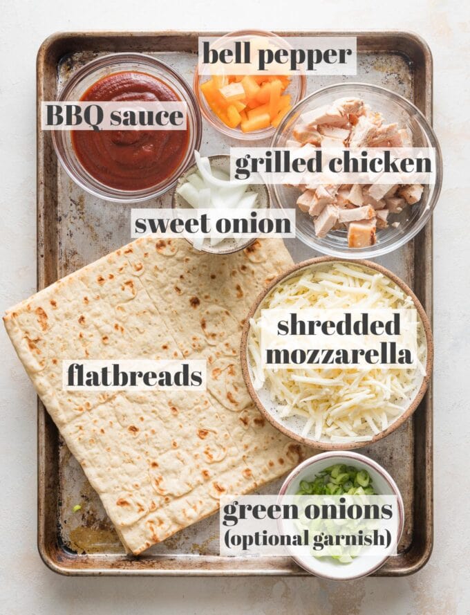 Overhead view of two flatbreads, BBQ sauce, shredded mozzarella, diced bell pepper, diced grilled chicken, sweet onion, and finely chopped green onions all in prep bowls and arranged on a sheet pan, ready to cook.