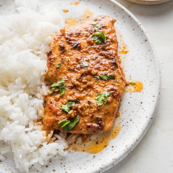 Cajun butter chicken breast with plenty of sauce, parsley for garnish, and a side of white rice on a white ceramic plate.
