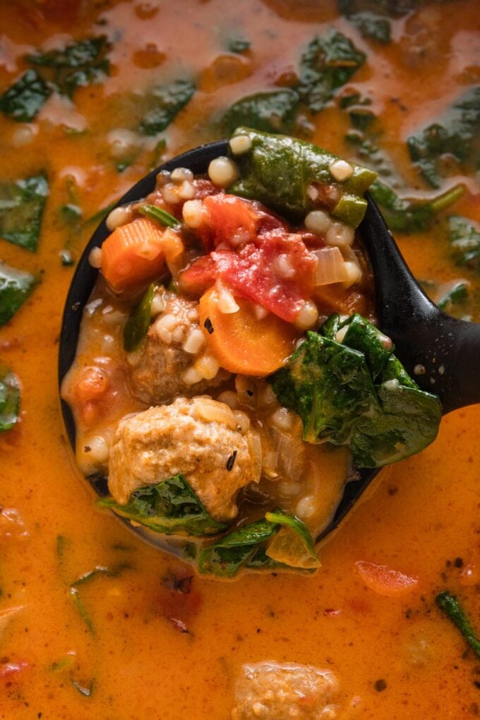 Close up image of a ladle lifting a portion of creamy tomato soup with pearl couscous and mini meatballs out of a Dutch oven.