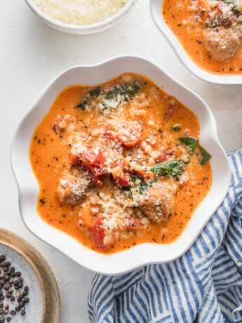 Small white bowls filled with creamy tomato meatball soup with extra Parmesan and black pepper in the background for garnish.