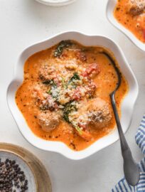 Small scalloped white bowl with a portion of creamy tomato meatball soup with pearl couscous and Parmesan.