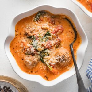 Small scalloped white bowl with a portion of creamy tomato meatball soup with pearl couscous and Parmesan.