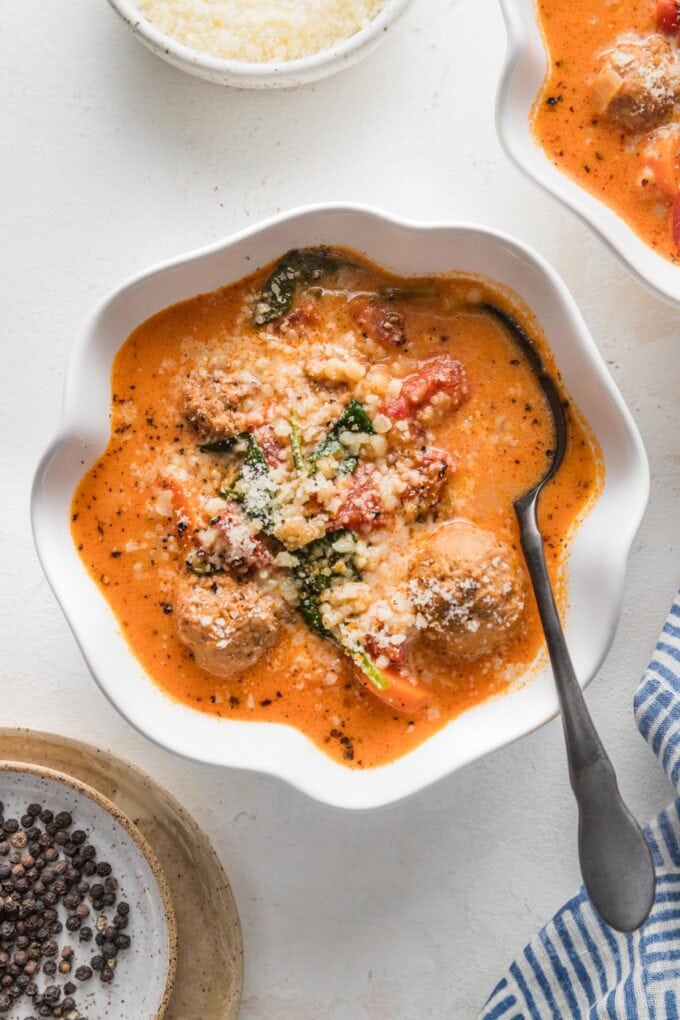 Small scalloped white bowl with a portion of creamy tomato meatball soup with pearl couscous and Parmesan.