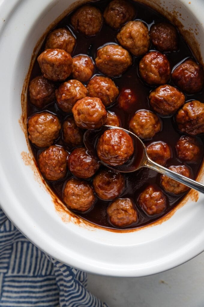 Spoon lifting a BBQ sauce and grape jelly-coated mini meatball up out of a small white slow cooker.