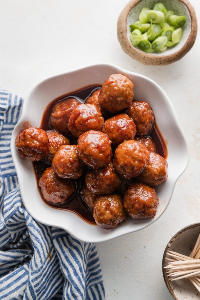 Bowl full of grape jelly BBQ meatballs cooked in the slow cooker and served as an appetizer with toothpicks and green onions nearby on the counter.