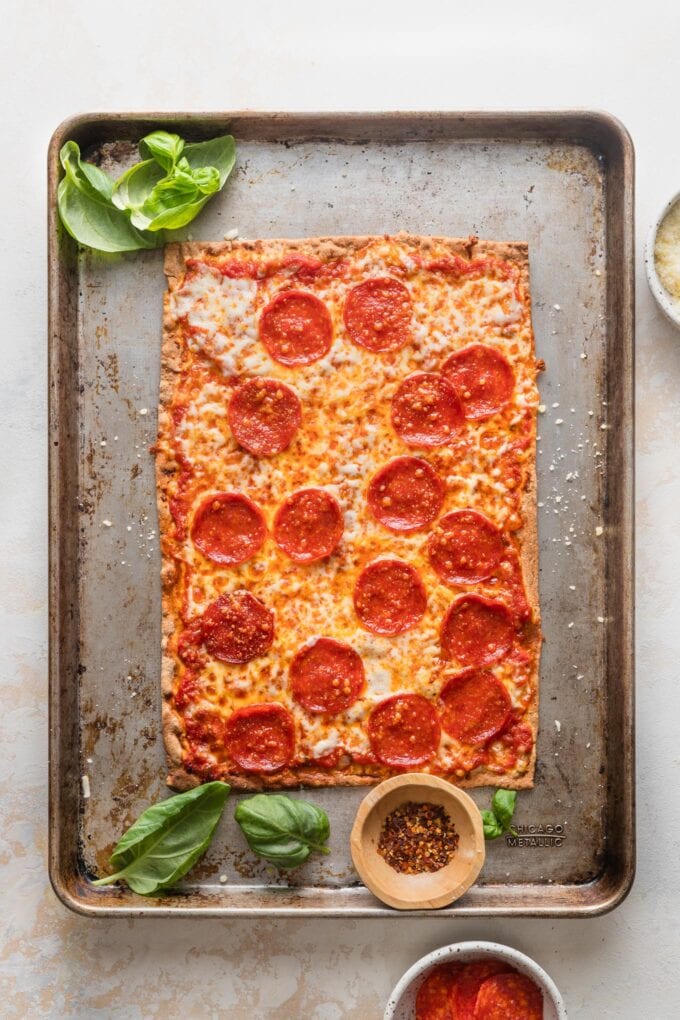 Sheet pan holding a flatbread-style pepperoni pizza and extra basil and red pepper to sprinkle on top.