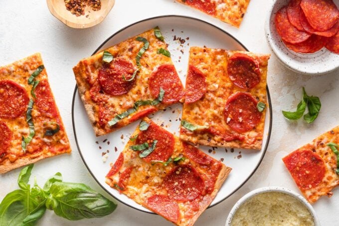 Small black-rimmed white plate with three pieces of pepperoni flatbread pizza, with extra pieces and fresh basil leaves scattered nearby..
