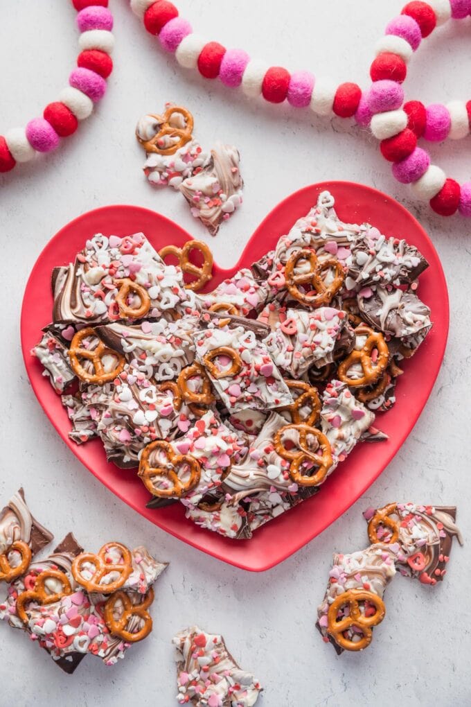 Red heart-shaped plate piled high with Valentine's-themed chocolate pretzel bark.