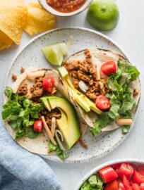 Two ground chicken tacos on a plate topped with avocado, green leaf lettuce, cheese, and tomato.