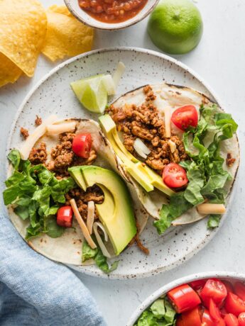 Two ground chicken tacos on a plate topped with avocado, green leaf lettuce, cheese, and tomato.