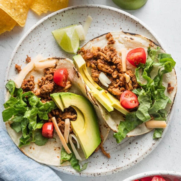 Two ground chicken tacos on a plate topped with avocado, green leaf lettuce, cheese, and tomato.
