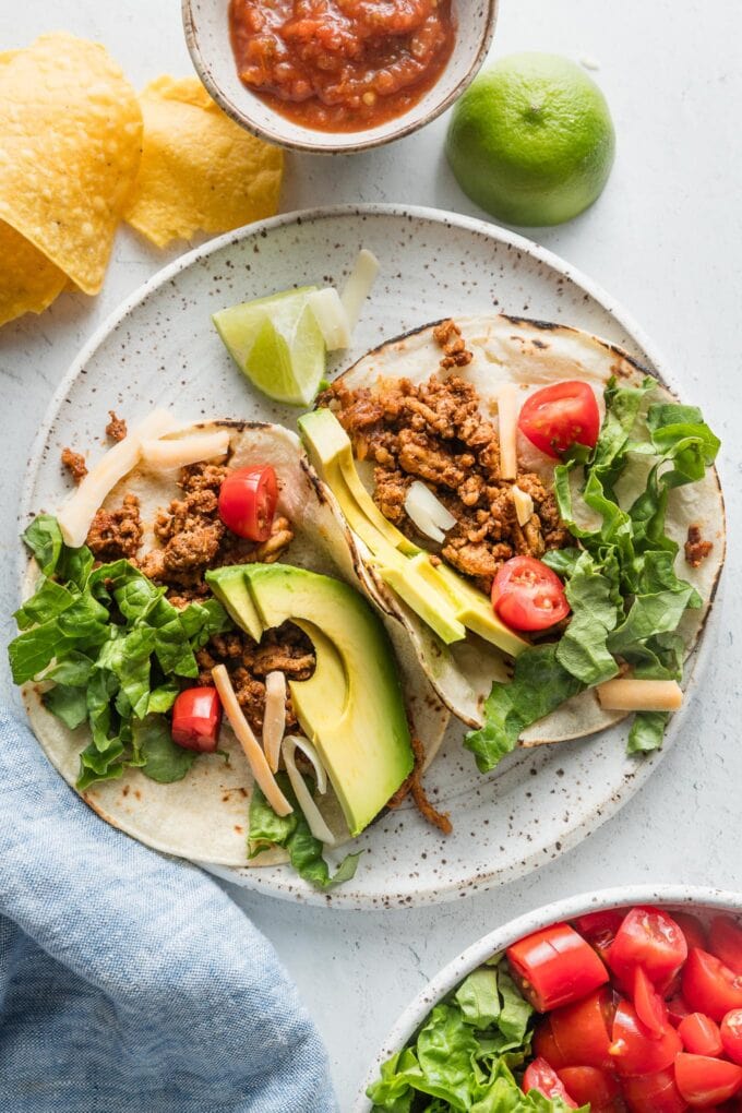 Two ground chicken tacos on a plate topped with avocado, green leaf lettuce, cheese, and tomato.