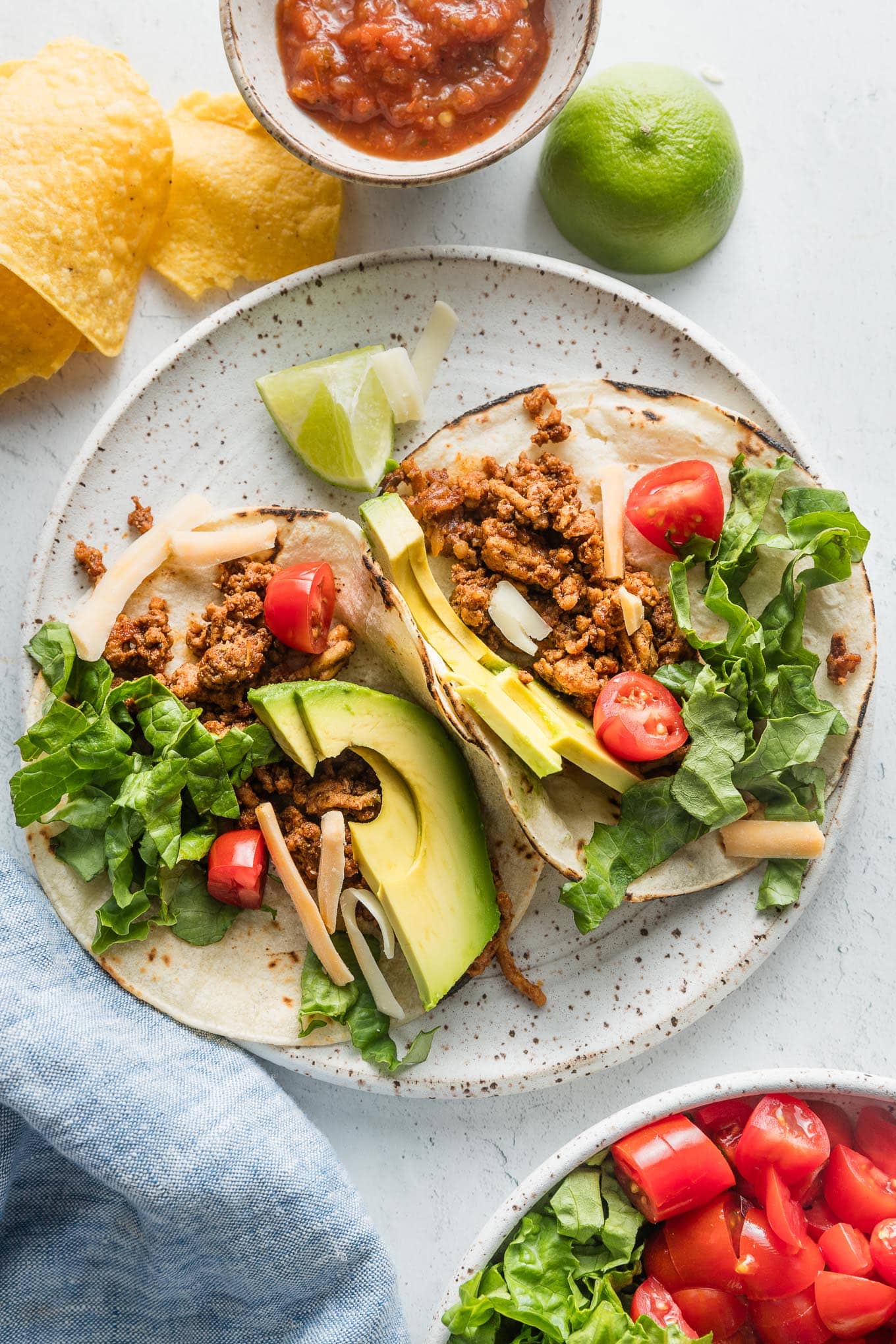 Two ground chicken tacos on a plate topped with avocado, green leaf lettuce, cheese, and tomato.