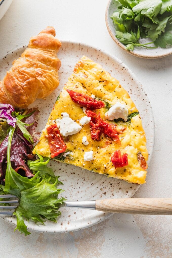 Slice of frittata topped with sun-dried tomatoes and feta, plated with a miniature croissant and salad greens.