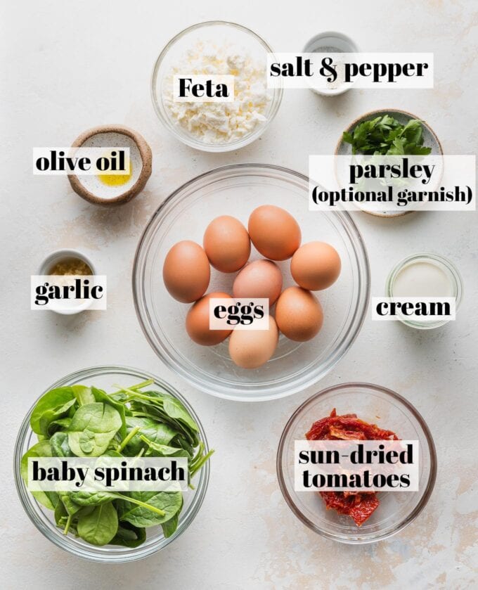 Labeled overhead photo of eggs, Feta, salt, pepper, cream, parsley, olive oil, garlic, baby spinach, and sun-dried tomatoes, all in prep bowls and ready to cook.