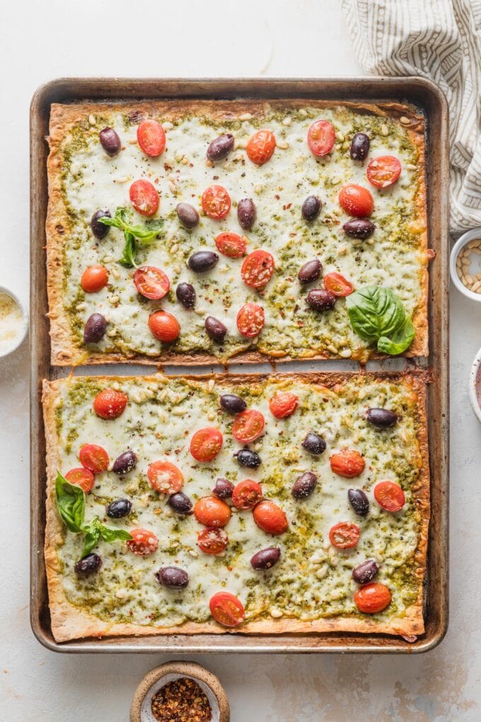 Baked pesto flatbread pizza with cherry tomatoes, olives, pine nuts, and fresh basil.