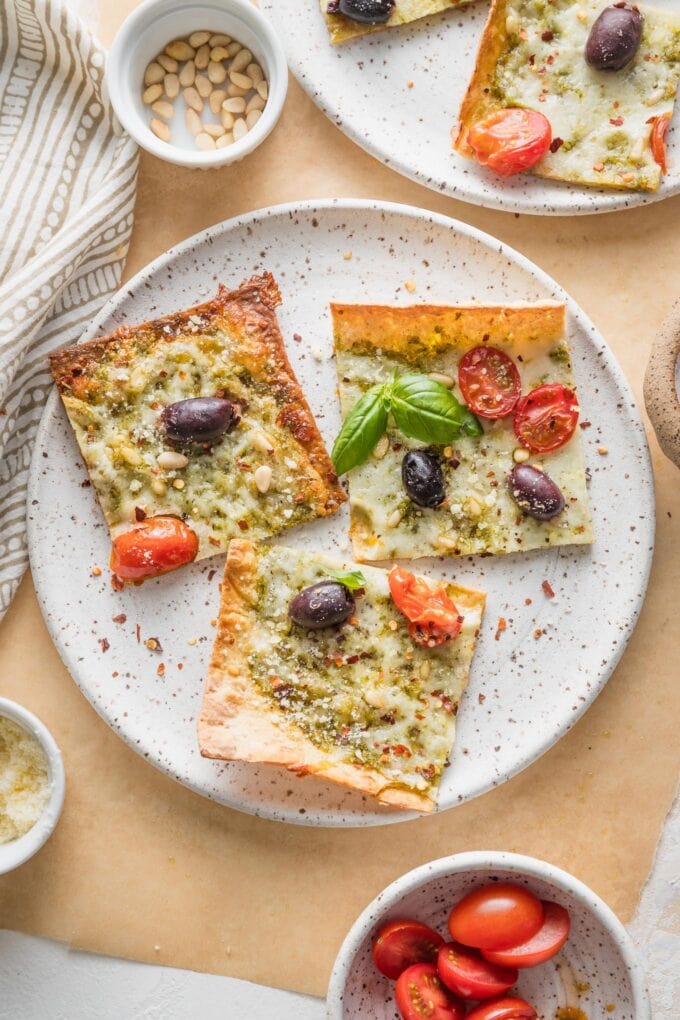 Parchment paper lined table with plates of pesto flatbread pizza sprinkled with fresh basil, pine nuts, and red pepper flakes.