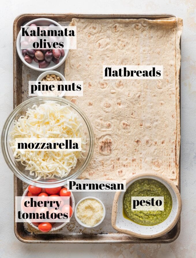 Labeled overhead photo of lavash style flatbreads, basil pesto, Kalamata olives, pine nuts, cherry tomatoes, mozzarella and Parmesan cheese, all in prep bowls arranged on a large rimmed baking sheet.
