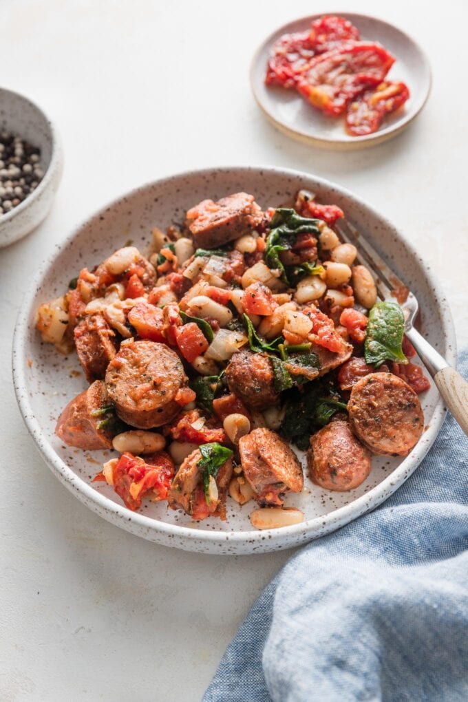 Angled view of a fork poised to lift a bite of Tuscan chicken sausage mixed with white beans, baby spinach, sun-dried and diced tomatoes, and Italian herbs.