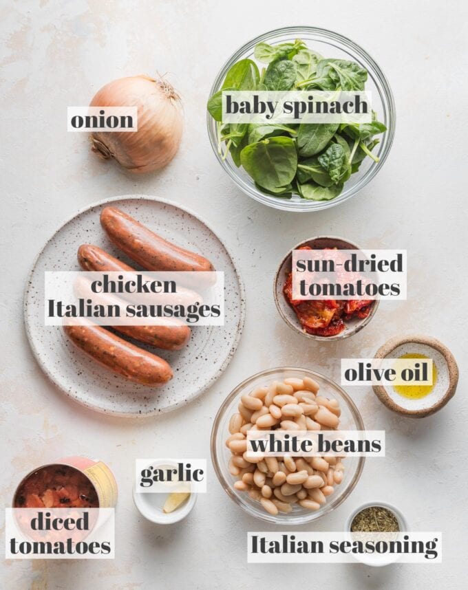 Labeled overhead photo of sweet Italian style chicken sausage links, baby spinach, canned white beans, sun-dried tomatoes, diced tomatoes, garlic, olive oil, and Italian seasoning, all measured into prep bowls and ready to cook.