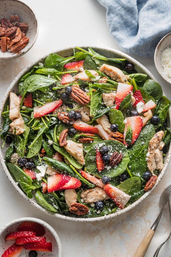 Large bowl full of berry pecan chicken spinach salad, with a blue napkin in the background as well as extra berries, pecans, and feta.