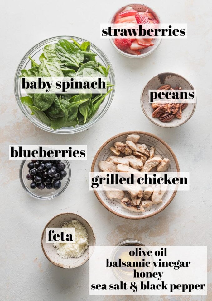 Labeled overhead photo of baby spinach, grilled chicken breast, strawberries, blueberries, pecans, feta, and salad dressing, all in prep bowls and ready to combine into salad.