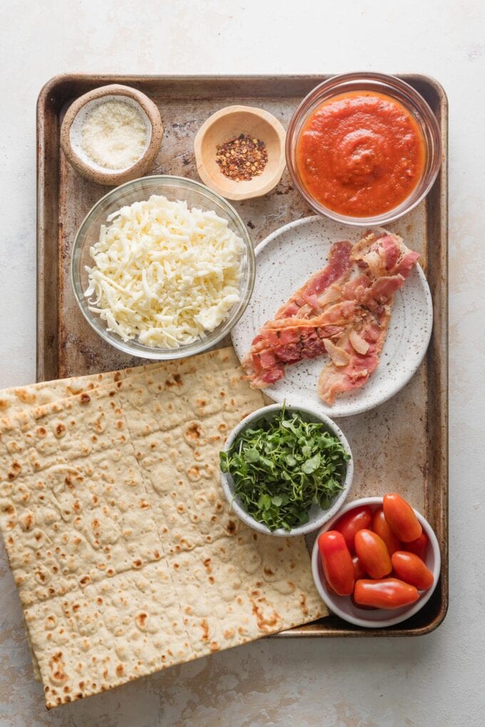 Baking sheet with flatbreads, marinara sauce, shredded mozzarella cheese, cooked bacon slices, tomatoes, and greens, ready to assemble into flatbread-style pizzas.
