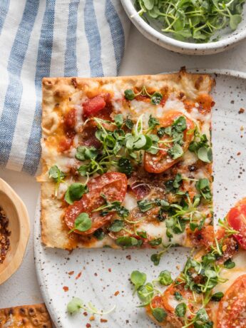 Close up of a BLT flatbread pizza slice on a plate with extra greens and red pepper flakes scattered around.