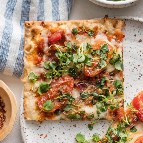 Close up of a BLT flatbread pizza slice on a plate with extra greens and red pepper flakes scattered around.