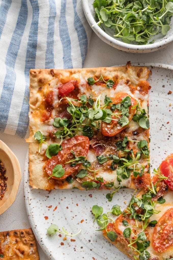 Close up of a BLT flatbread pizza slice on a plate with extra greens and red pepper flakes scattered around.