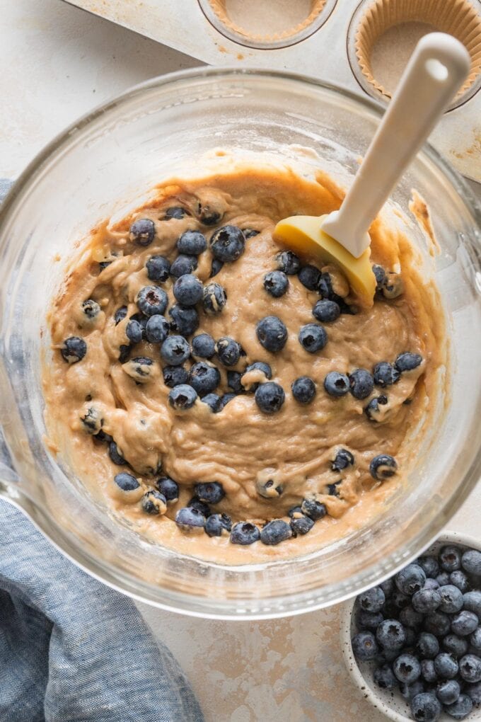 Close-up of a thick banana muffin batter with blueberries mixed in, in a clear glass mixing bowl.