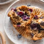 Small white speckled ceramic plate with a blueberry banana muffin pulled apart to show the moist interior and crunchy top.