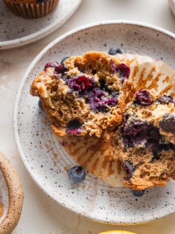 Small white speckled ceramic plate with a blueberry banana muffin pulled apart to show the moist interior and crunchy top.