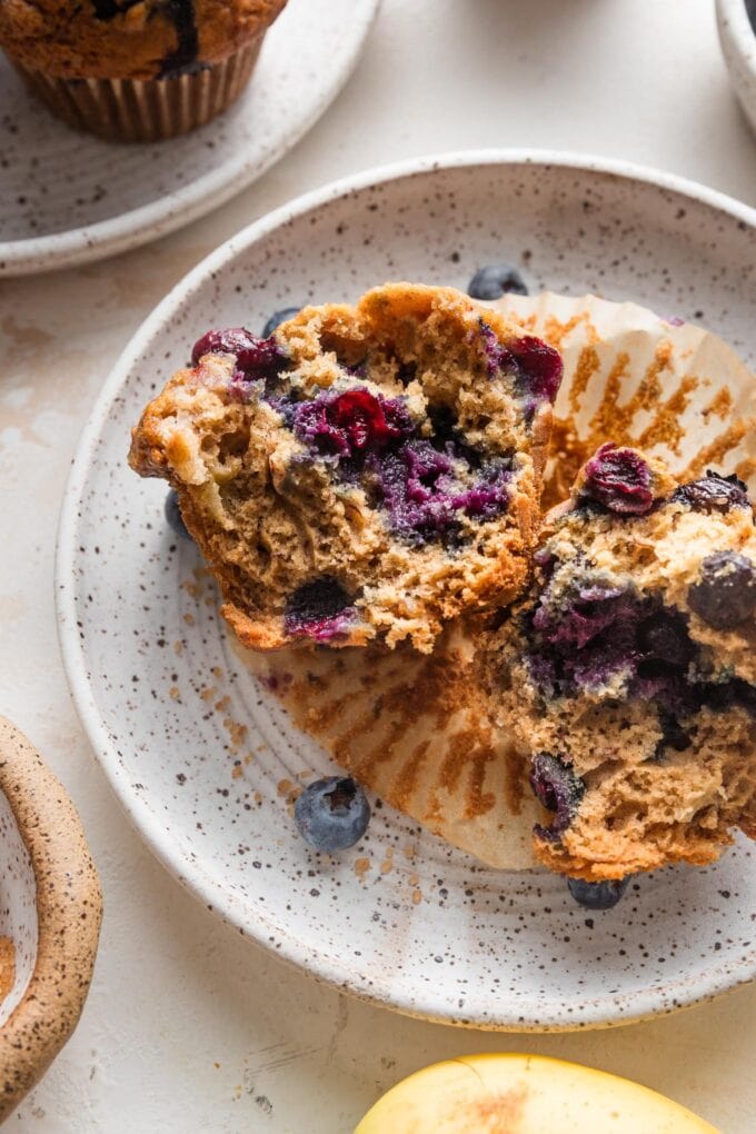 Small white speckled ceramic plate with a blueberry banana muffin pulled apart to show the moist interior and crunchy top.