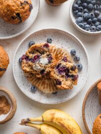 Countertop scattered with banana blueberry muffins, extra bananas and blueberries, and coarse sparkling sugar.