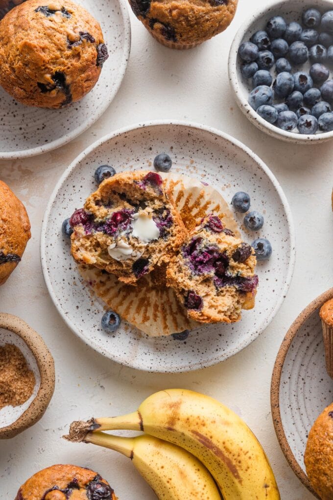 Countertop scattered with banana blueberry muffins, extra bananas and blueberries, and coarse sparkling sugar.