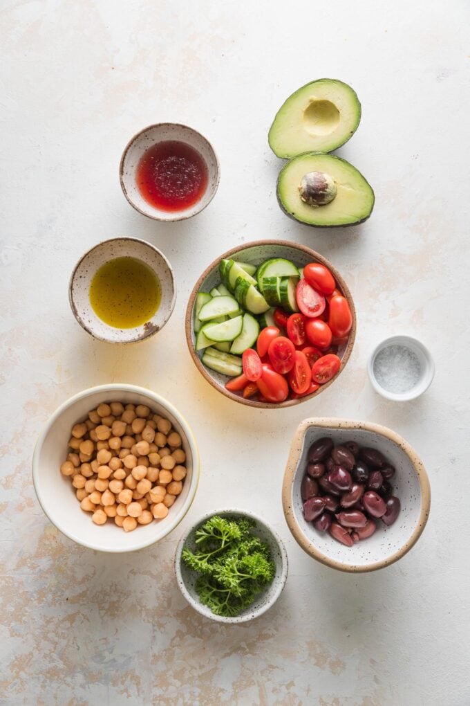 Overhead photo of canned chickpeas, drained and rinsed, chopped cucumber and tomatoes, a halved avocado, Kalamata olives, fresh parsley, olive oil, red wine vinegar, and kosher salt, all in prep bowls and ready to combine into a salad.