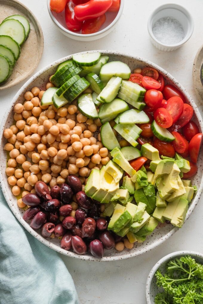 Low bowl full of chickpeas, cucumbers, olives, and avocado, in sections and not yet mixed together.