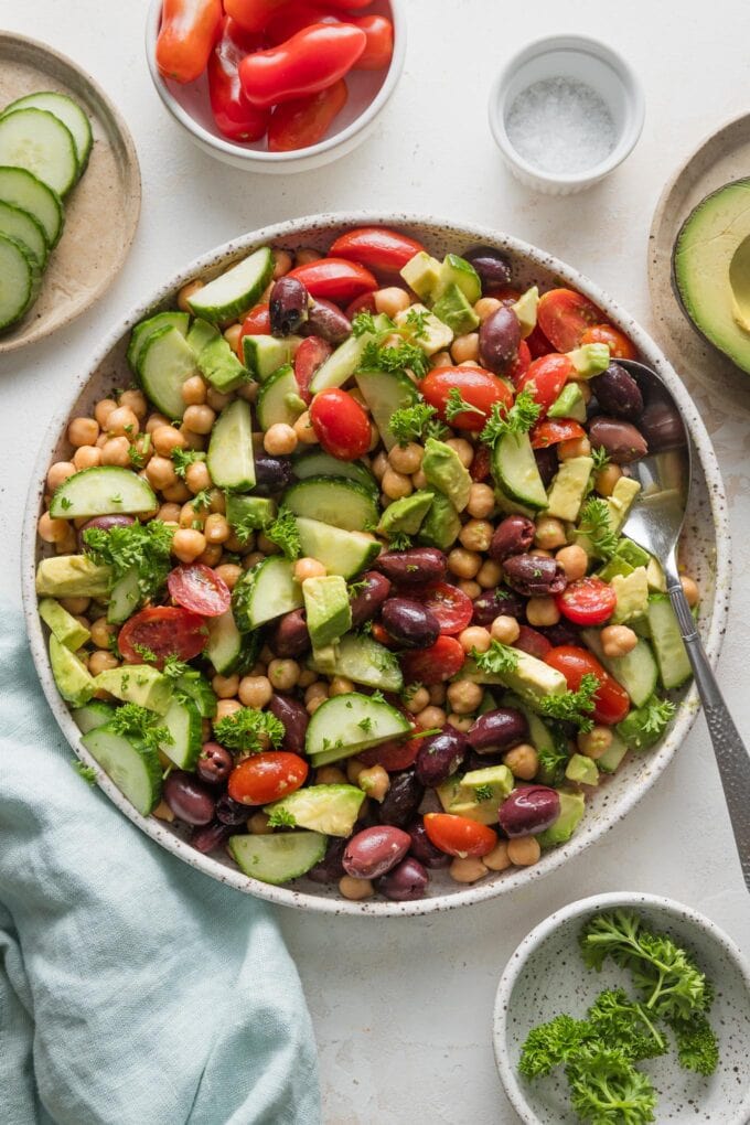 Countertop with a bowl full of chickpea avocado salad in focus, with extra prep bowls of veggies and a linen kitchen towel in the background.