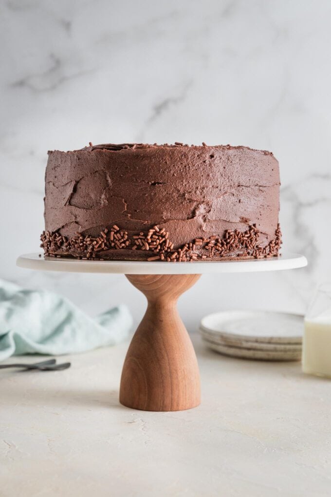 Homemade chocolate layer cake with chocolate buttercream frosting, whole and ready to serve on a white and wooden cake stand.