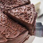 Slice of homemade buttermilk chocolate layer cake being lifted off the cake plate to show the creamy chocolate frosting and moist, rich chocolate interior.