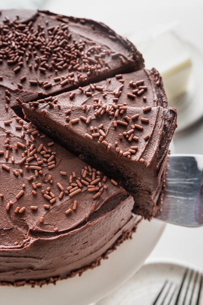 Slice of homemade buttermilk chocolate layer cake being lifted off the cake plate to show the creamy chocolate frosting and moist, rich chocolate interior.