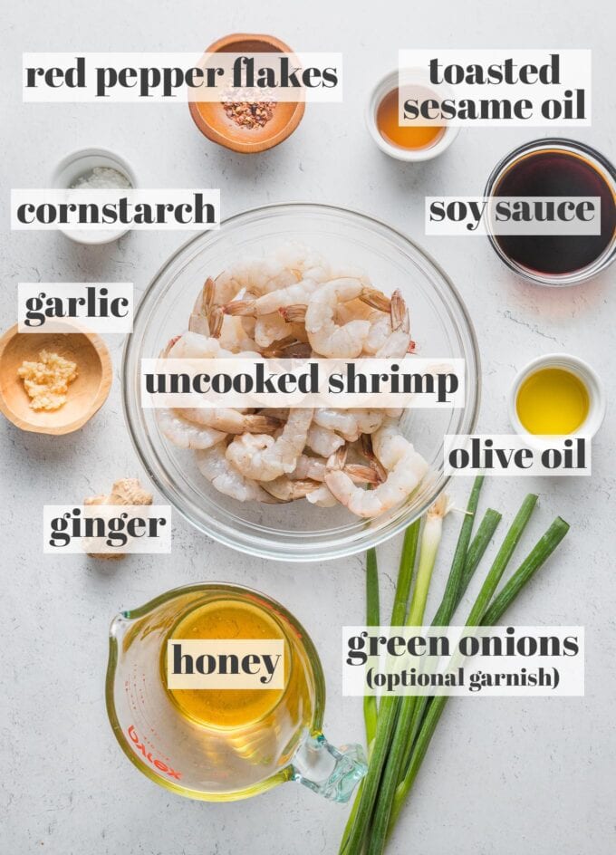 Labeled overhead photo of raw shrimp, honey, soy sauce, toasted sesame oil, olive oil, garlic, ginger, red pepper flakes, cornstarch, and green onions, laid out on a countertop and ready to cook.
