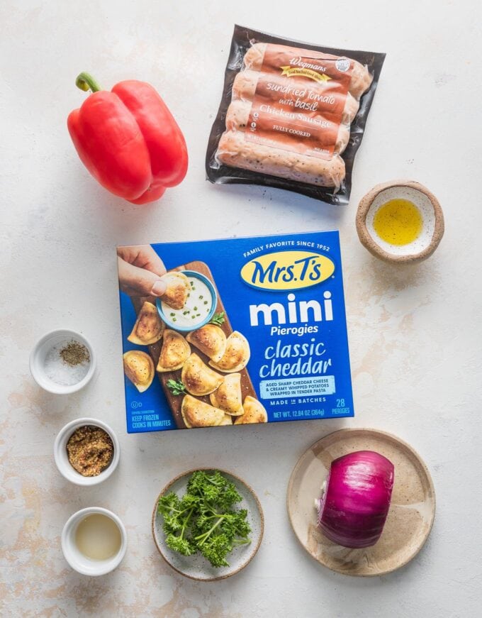 Overhead view of a box of Mrs. T's mini pierogies, fully cooked chicken sausage links, a red bell pepper, a red onion, olive oil, whole grain mustard, apple cider vinegar, salt, pepper, and fresh Italian parsley, all ready to cook in a sheet pan meal.