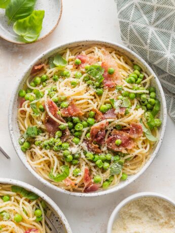 Low bowl filled with pasta with bacon, peas, and a light emulsified olive oil-based sauce, topped with Parmesan, fresh basil, and black pepper.