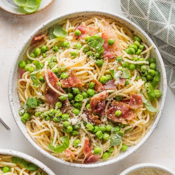 Low bowl filled with pasta with bacon, peas, and a light emulsified olive oil-based sauce, topped with Parmesan, fresh basil, and black pepper.